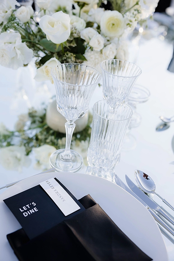 Reception tablescape featuring a black and white tablescape with white roses and greenery, crystal glasses, silver flatware, and menu card on plate