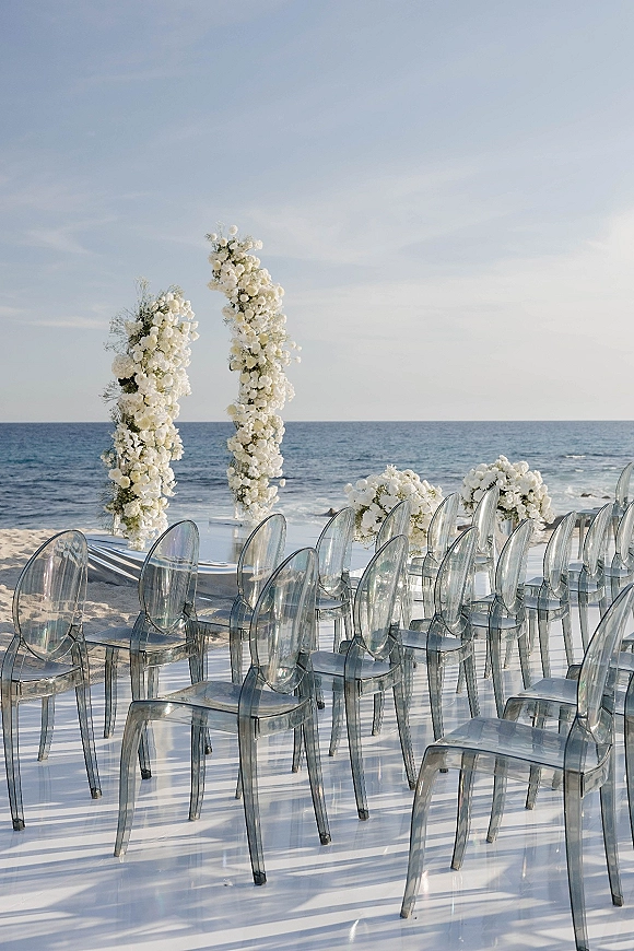 Beach ceremony setup with a white floral arch, aisle runner, and clear acrylic chairs on sand overlooking the ocean horizon
