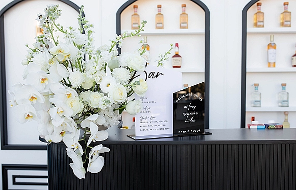 Wedding bar decor featuring a wedding bar sign and white orchid, rose, and hydrangea arrangement on a bar counter before arched shelves of bottles