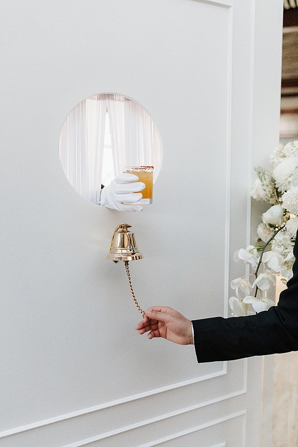 Wedding cocktail wall with bell pull and gloved hand serving a drink through a round window, framed by white panels and sheer curtains