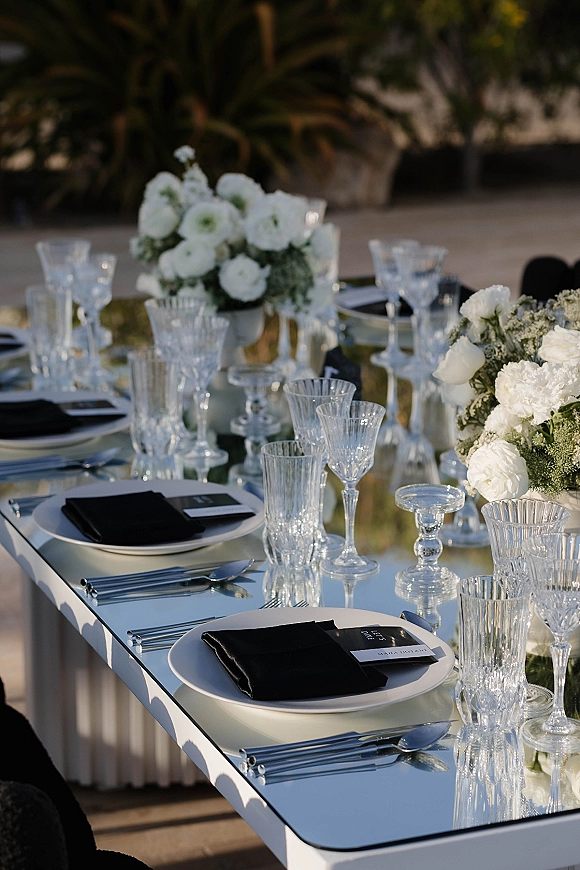 Reception tablescape with black and white wedding table styling, white flower centerpieces, crystal stemware, and clear votives in a garden setting