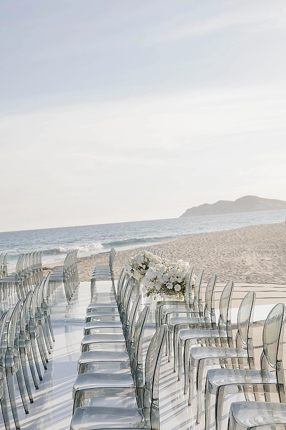 Beach ceremony setup with oceanfront wedding ceremony aisle, clear chairs lining a white runner with white florals on sandy shore