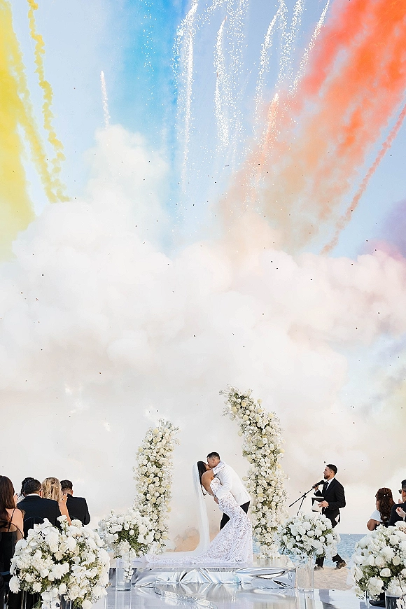 Ceremony kiss moment as newlyweds dip kiss on an oceanfront altar with white floral pillars, veil, and fireworks with colored smoke overhead