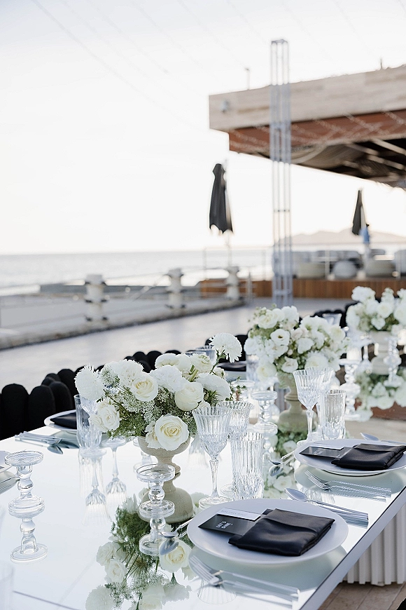 Reception tablescape with black and white tablescape details, mirrored tabletop, white rose centerpieces, crystal goblets, and oceanfront patio backdrop