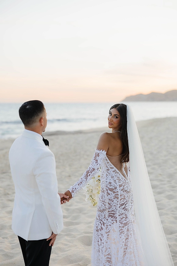 Couple portrait holding hands on a sandy beach at sunset, bride looking back in lace dress with long veil and bouquet beside groom in white tuxedo