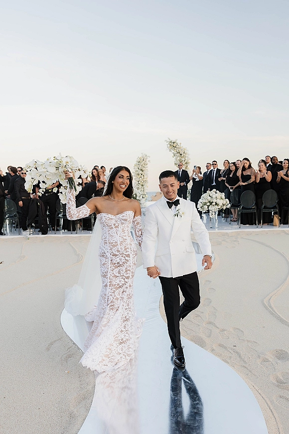 Wedding recessional as bride and groom walk down aisle hand in hand, bride raising bouquet beneath white arch on beach by ocean horizon