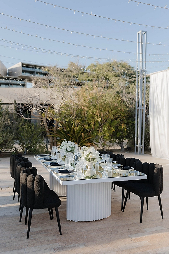 Reception tablescape with outdoor reception tablescape featuring a mirrored long table, black chairs, white florals, goblets, and string lights on a patio