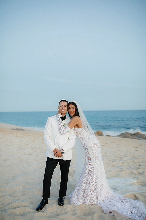 Couple portrait on a sandy beach, bride in lace dress with veil hugging groom in tuxedo with boutonniere, ocean and rocks behind