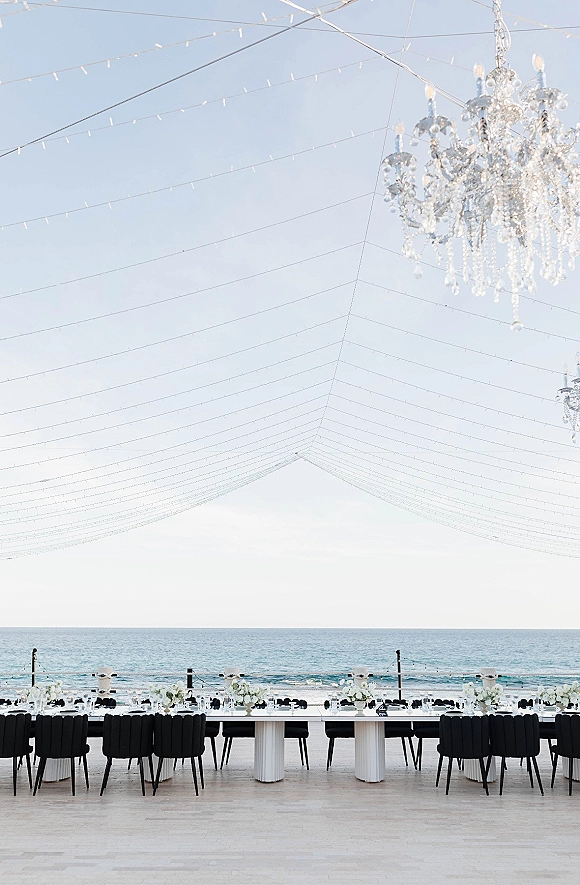Reception tablescape with a long banquet table wedding setup, white florals and candles under string lights on a deck with ocean view