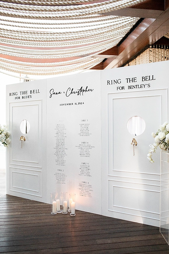Wedding seating chart seating chart wall with white escort panels, bell pull chains, and candle cylinders on a wooden deck under a rope canopy