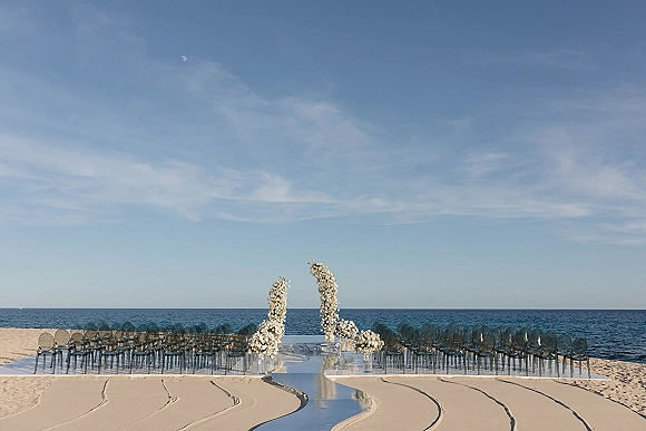 Beach ceremony setup with mirrored aisle, clear chairs, and white floral arches on a sandy shore with ocean horizon under blue sky