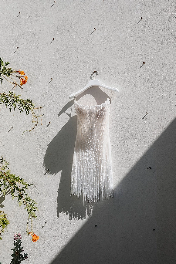 Wedding dress on a white hanger with beaded fringe and spaghetti straps, hanging on a sunlit stucco wall with greenery and orange flowers