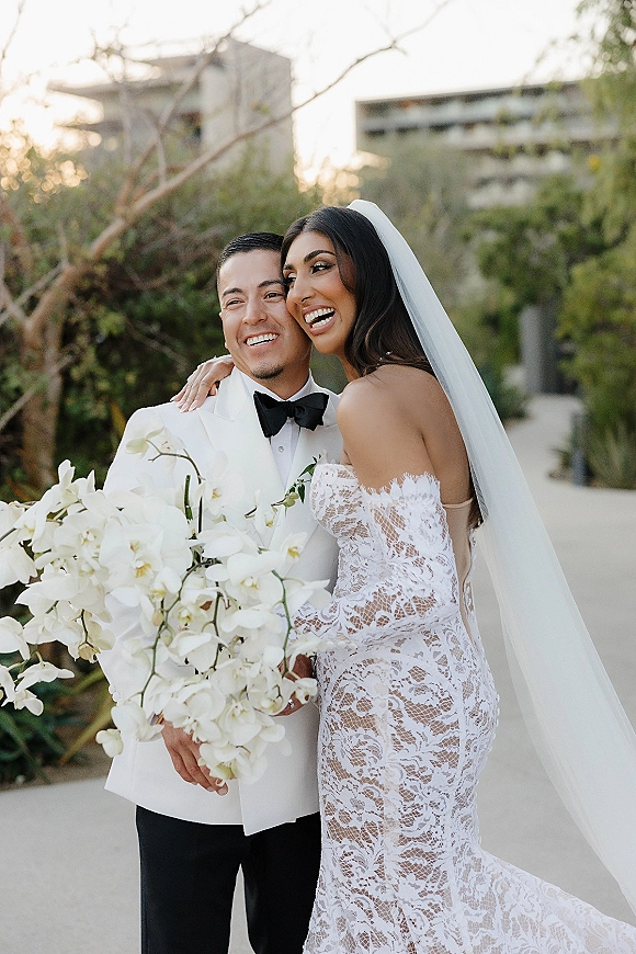 Couple portrait of bride and groom laughing as she hugs him, lace gown and veil flowing, holding orchid bouquet on a green walkway near a modern building