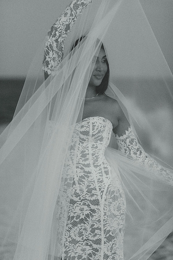 Bridal portrait in black and white of a bride under veil, wearing a strapless lace corset gown, lace gloves, and necklace outdoors near horizon