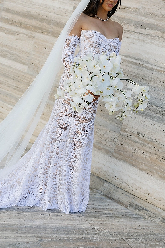 Bridal portrait of a bride in a strapless lace wedding dress holding a white orchid bouquet with a long veil against a stone wall backdrop