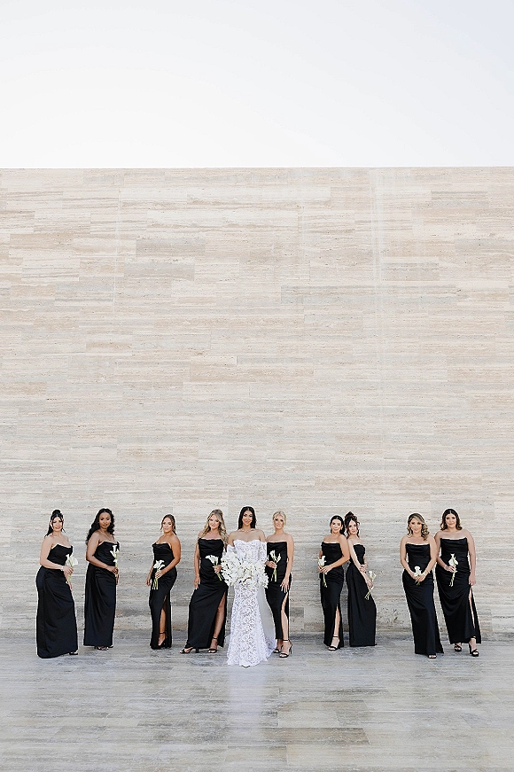 Bridesmaid group photo of bride with bridesmaids in strapless black dresses holding white calla lilies against a neutral stone wall