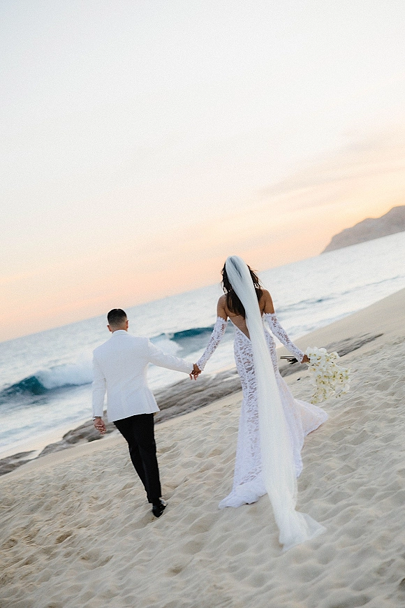 Couple portrait of newlyweds holding hands walking away on a beach at sunset, bride’s veil trailing, bouquet and ocean waves behind