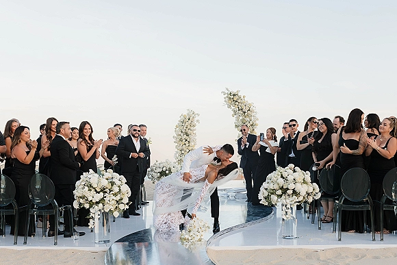 Wedding kiss as the bride and groom dip at a white floral arch, her veil flowing, with guests clapping by the ocean horizon
