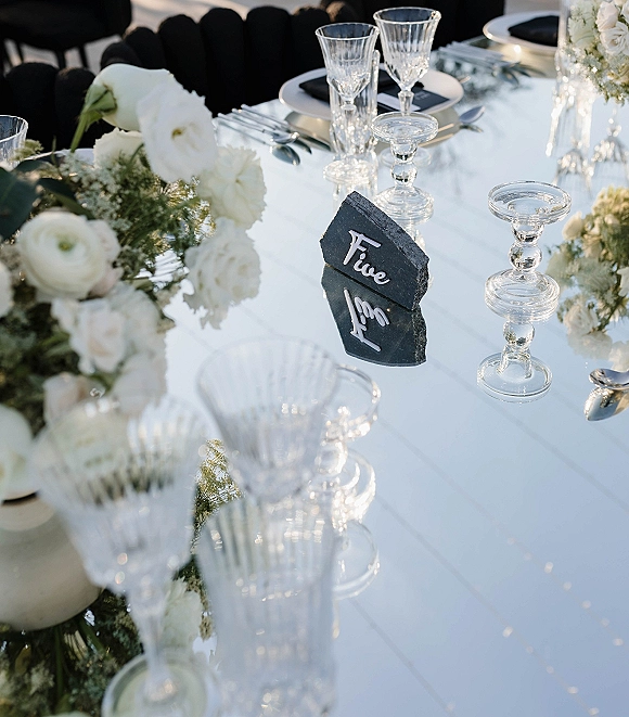 Reception tablescape with mirrored wedding table, white floral centerpiece and greenery, crystal wine glasses, black plates, slate number in outdoor light