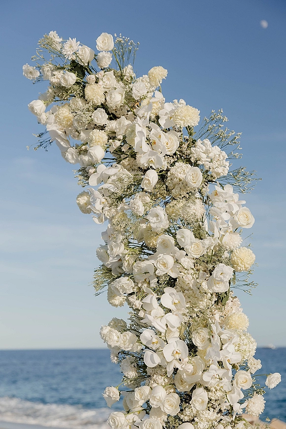 Ceremony floral arch of white roses and orchids with baby's breath and greenery, framed against an ocean horizon and blue sky