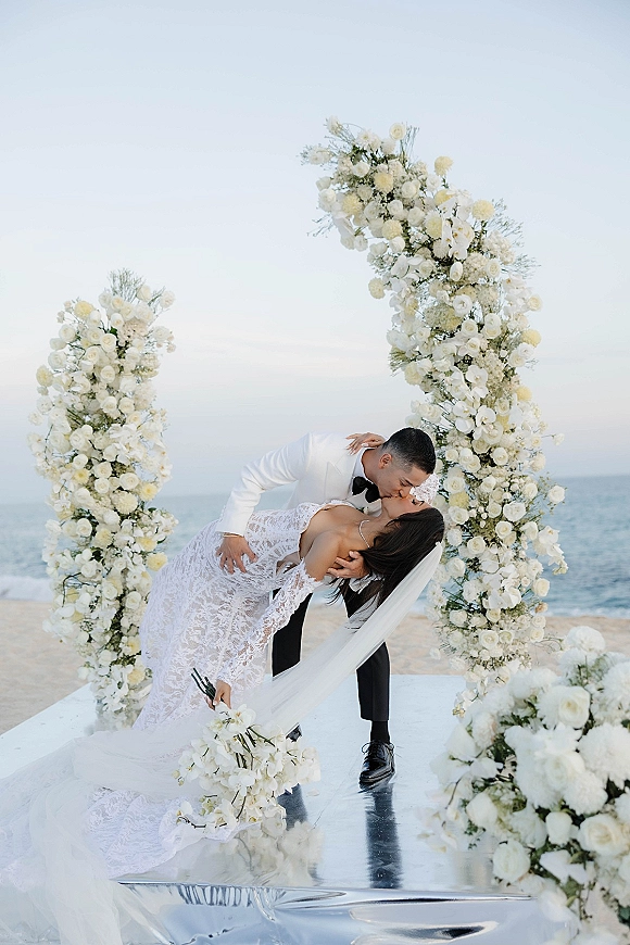 Wedding kiss portrait of bride and groom in a dip kiss wedding photo under a white floral arch on the beach, veil blowing by the ocean