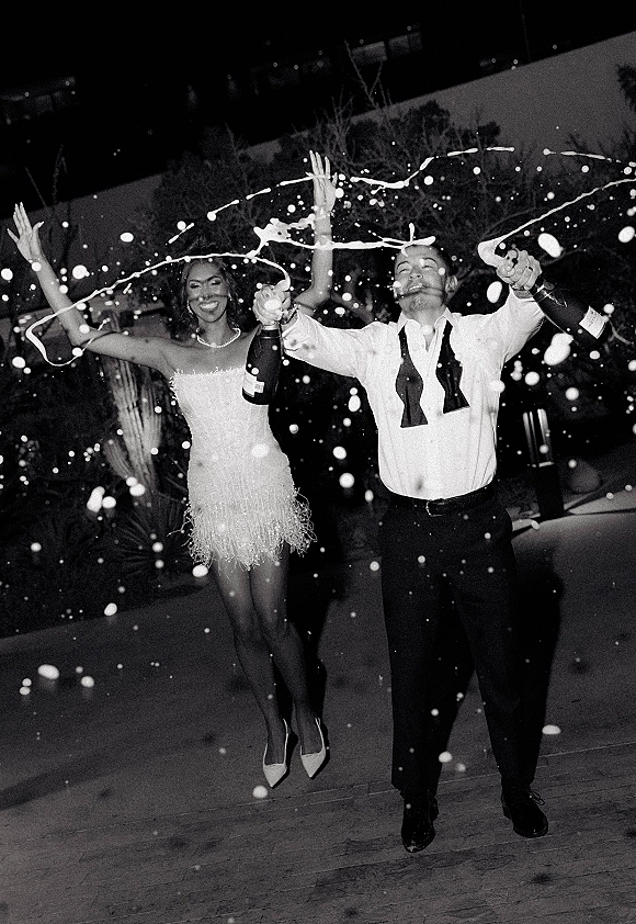 Champagne pop moment as bride in beaded mini dress and pearl necklace and groom with untied bow tie spray bottles on patio at night