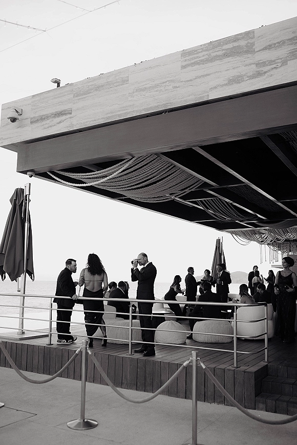 Wedding cocktail hour with guests mingling in a wedding lounge area, holding cocktails under a string canopy on a deck with ocean views