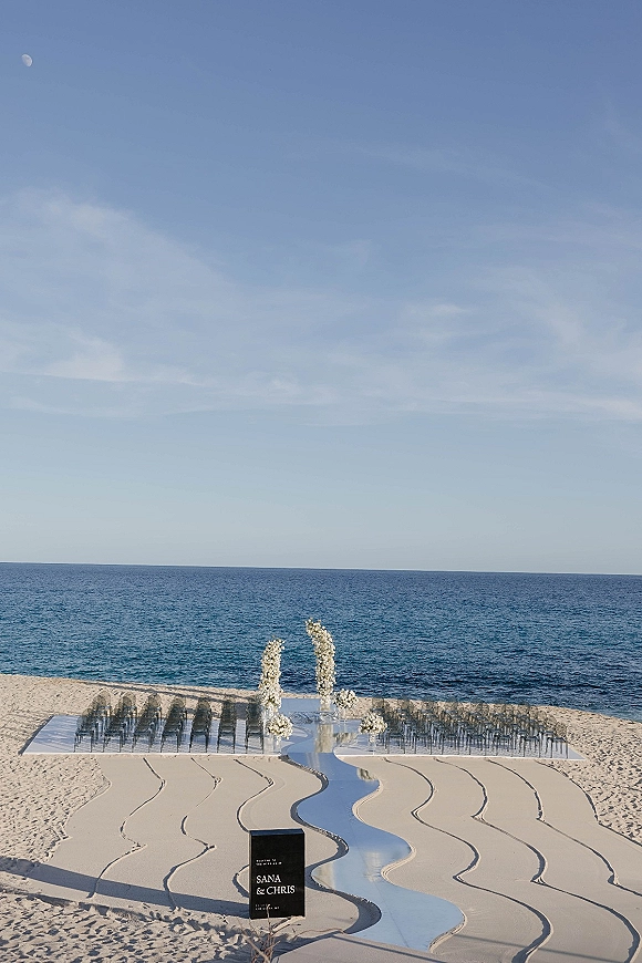 Beach ceremony setup with clear chairs lining a mirrored aisle runner to a white floral arch on sand, ocean horizon behind