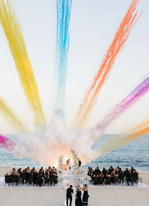 Ceremony kiss moment as the bride and groom kiss beneath a white floral arch, smoke bombs drifting over an oceanfront beach aisle