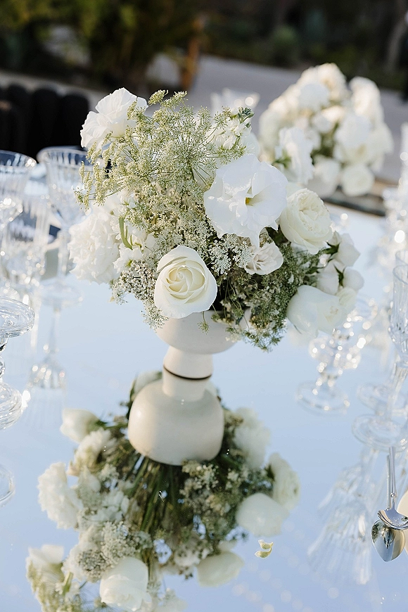 Wedding centerpiece with a white floral centerpiece of roses and filler blooms in a ceramic vase on a white tablecloth with glassware, silverware, greenery beyond