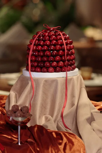Dessert tower anchors a wedding dessert display, wrapped with a red ribbon at an indoor reception framed by lush greenery.