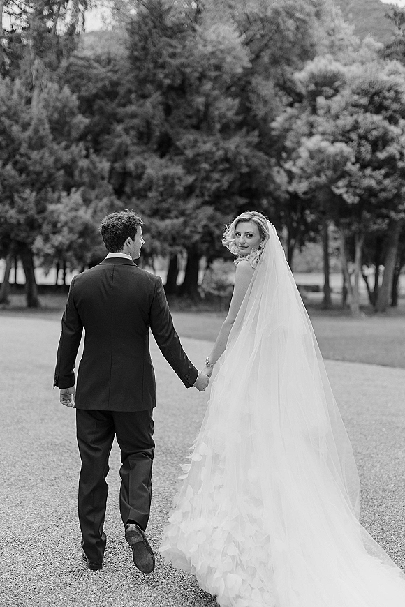 Couple portrait of bride and groom holding hands, walking away on a tree-lined gravel path as her veil trails behind