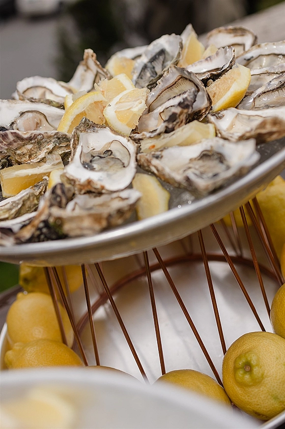 Oyster bar display with oysters on the half shell on ice and lemon wedges on a metal tray stand at an outdoor greenery catering station
