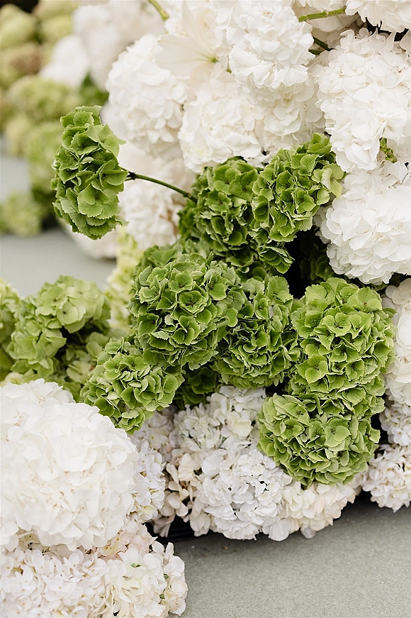 Hydrangea arrangement of white and green blooms with stems, styled as a wedding centerpiece on a stone surface with blurred florals behind