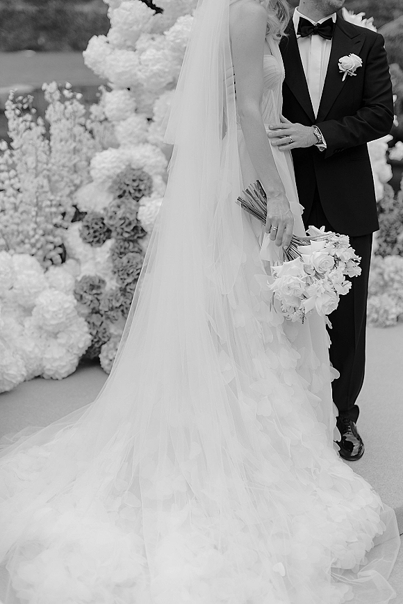 Wedding couple portrait, black and white wedding portrait of bride with bouquet and long veil beside groom in tuxedo by a flower wall