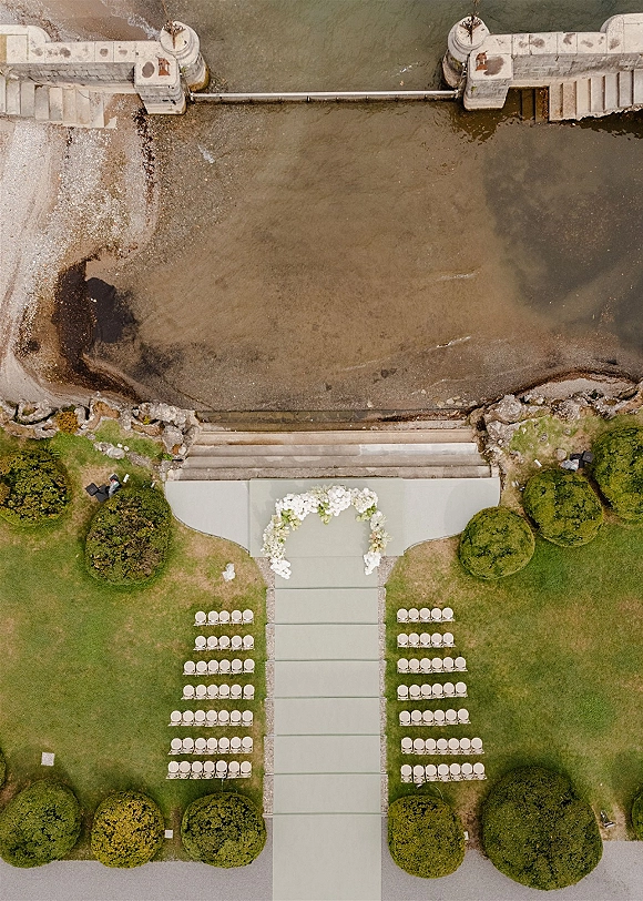 Outdoor ceremony setup with a white floral arch and aisle runner, chairs on a green lawn facing the shoreline and water beyond