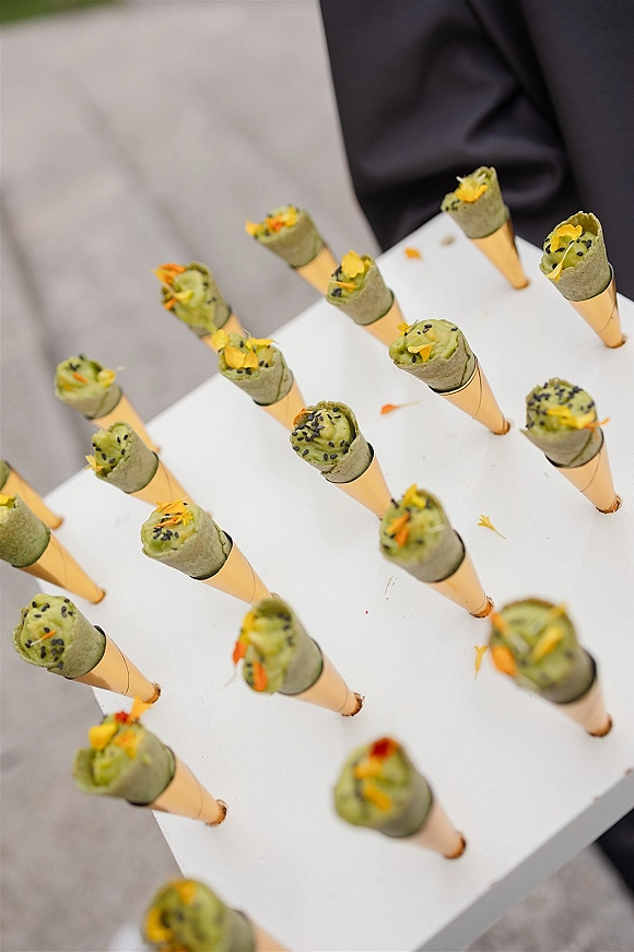 Cocktail hour appetizers in paper cones with herb puree, black sesame, and edible petals arranged on a board on a white tabletop