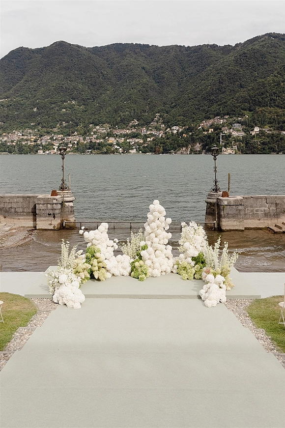 Ceremony backdrop with outdoor wedding altar of white hydrangeas and green florals on a platform beside a lake and mountain hillside