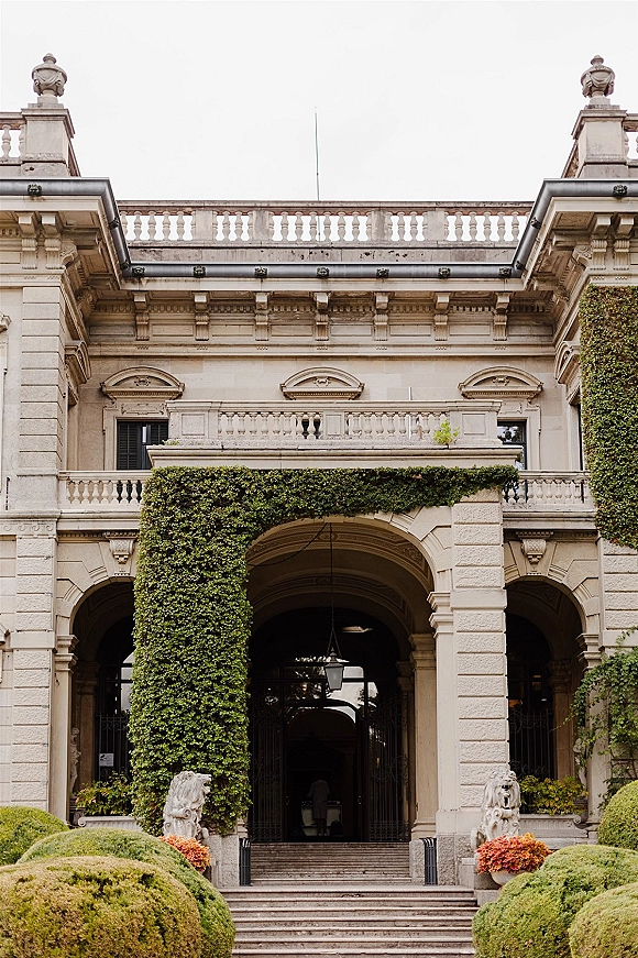 Wedding venue exterior with an ivy-covered archway and stone stairs, framed by lion statues and wrought iron gates on a grand facade