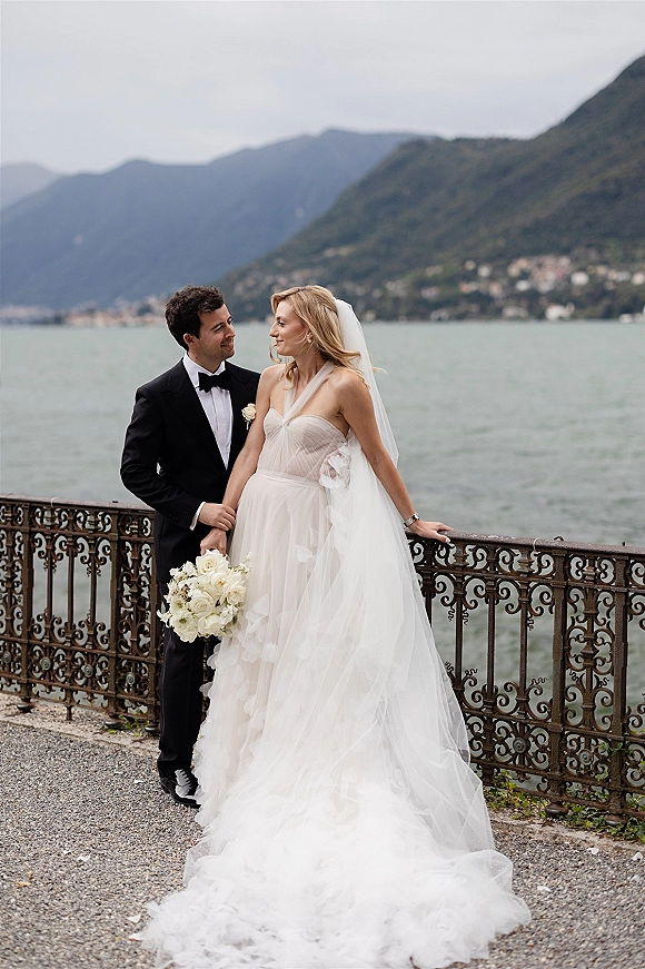Couple portrait of bride and groom by lake, gazing at each other as she leans on wrought iron railing with veil and bouquet, mountains behind