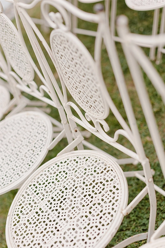 Wedding ceremony chairs in white metal line a grass lawn, showing lattice seats and wrought iron backs amid outdoor greenery