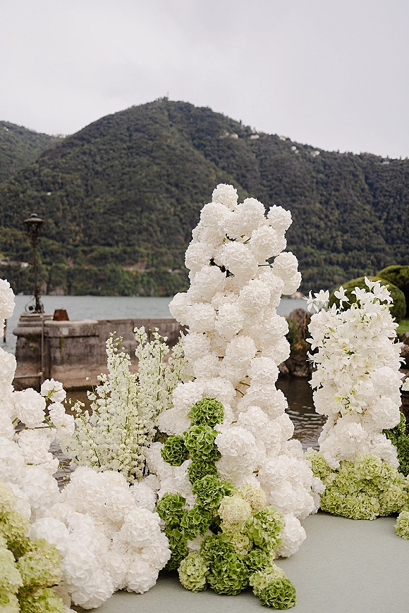 Wedding ceremony florals with a ceremony floral installation of white blooms and green hydrangeas on a stone terrace by a lake and mountains