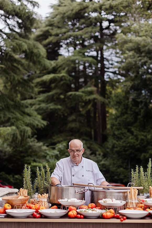Wedding catering station with chef beside stainless steel stock pots, soup bowls, breadsticks and rosemary planters set among trees outdoors