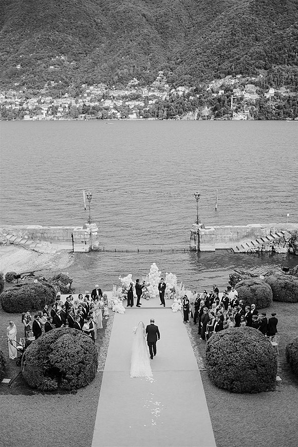 Outdoor wedding ceremony with bride walking down a garden ceremony aisle on a white runner, long veil flowing toward a lakeside altar
