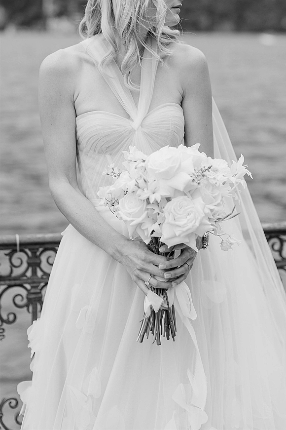 Bridal portrait of a bride holding bouquet of white roses with ribbon wrap, wearing a tulle halter gown and veil on a lawn by a wrought iron fence