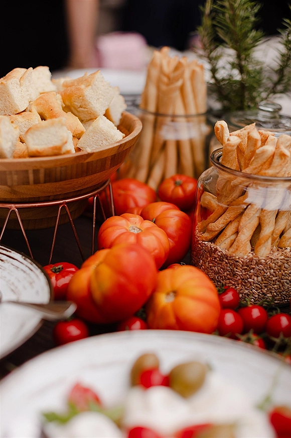 Wedding appetizer table with wedding cocktail hour snacks, bread cubes and breadsticks in a jar beside tomatoes and rosemary, guests blurred behind