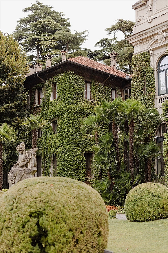 Wedding venue exterior with ivy-covered walls, arched windows, and terracotta roof, framed by palm trees and manicured lawn gardens