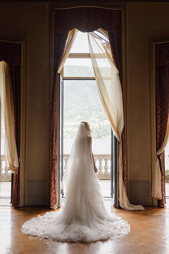 Bridal portrait of bride from behind in a strapless gown with cathedral veil and tulle train framed by open French doors, lake and mountains beyond