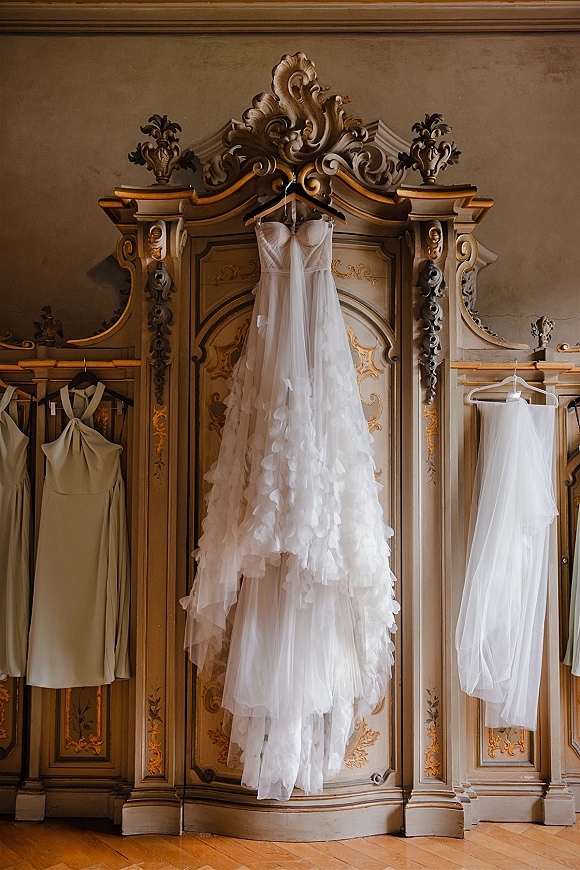 Wedding dress hanging on a hanger, strapless bridal gown with layered tulle skirt and long veil beside a bridesmaid dress on ornate wardrobe