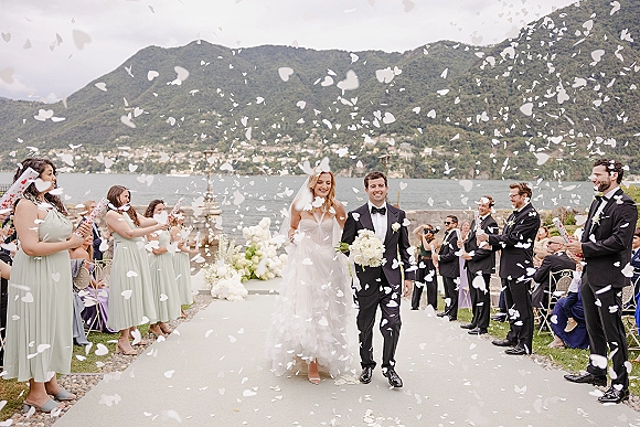 Wedding recessional as bride and groom walk the aisle under a confetti toss, her veil flowing on a lakeside terrace with mountains behind